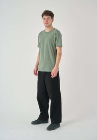 Grünes Baumwoll-T-Shirt mit Rundhalsausschnitt und kurzen Ärmeln, kombiniert mit lockeren schwarzen Hosen und dunkelgrünen Schuhen, stehend vor einem neutralen Hintergrund.