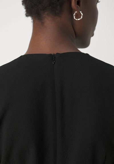 Vestido negro de tela texturizada, con escote recto en la espalda y un pequeño cierre de cremallera en la parte superior. Pendientes de aro plateados visibles.
