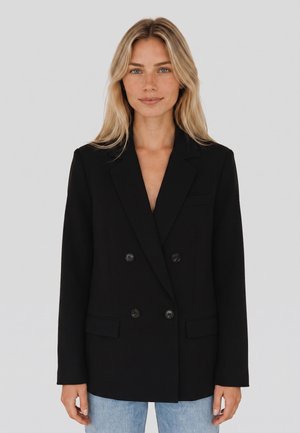 Donna con lunghi capelli biondi indossa un blazer nero doppiopetto e jeans azzurri chiari su uno sfondo grigio chiaro uniforme.