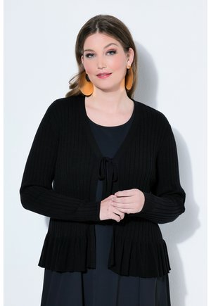 Vrouw met een lichte huid en bruin haar draagt grote oranje oorbellen, een zwarte geribbelde cardigan met strik aan de voorkant en een zwarte jurk, staand tegen een witte muur.