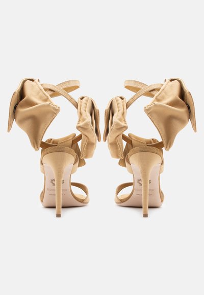 Ζευγάρι μπεζ πέδιλα με ψηλό τακούνι, διακοσμημένα με υφασμάτινα βολάν και λουριά στον αστράγαλο, εμφανιζόμενα από πίσω σε λευκό φόντο.