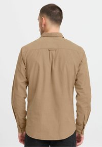 Camisa marrón de manga larga con cuello abotonado, que presenta un pliegue en la parte trasera y puños abotonados. Fabricada con un tejido ligero y de textura suave.