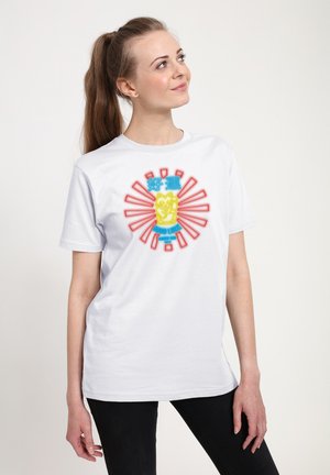 Kvinde med brunt hestehale iført hvid T-shirt med neongul lykkekat og rød solstråle-design, ser opad, står foran en hvid baggrund.