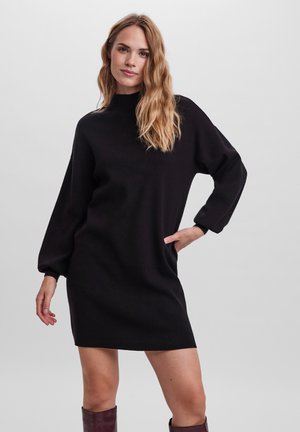 Robe noire en tricot avec un col montant, des manches ballons longues et des poches latérales, présentant une texture douce et une coupe décontractée.
