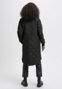 Abrigo acolchado largo negro con cintura ceñida, puños en las muñecas y dobladillo redondeado, usado sobre pantalones y botas de cuero negro.