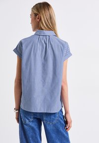 Camicia a maniche corte blu e bianca a righe con colletto, caratterizzata da un design con pieghe sulla parte posteriore e una texture di tessuto morbido. Abbinata a pantaloni in denim.