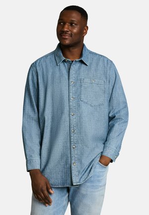 Man die een lichtblauw geruit denimoverhemd en lichtblauwe jeans draagt, kijkt opzij met één hand in de zak en de andere ontspannen.