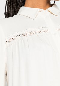 Krämfärgad skjortblus med krage, med spetstillägg och veckad textur framtill. Mjuk tyg och avslappnad passform.