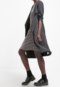 Žena jde oblečená v šatech přes kolena s kostkovaným vzorem houndstooth, dlouhém tmavém kabátu, černých ponožkách a černých botách na přezku.