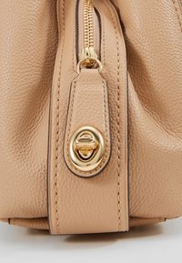 Sac à main en cuir beige avec une finition texturée, une fermeture éclair dorée et un accent de fermoir rond doré. La conception comprend des détails de couture subtils.