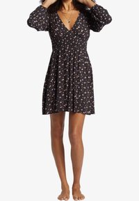 Žena v černých květovaných šatech s dlouhými rukávy a výstřihem do V, stojící bosá s rukama u hlavy, na bílém pozadí.