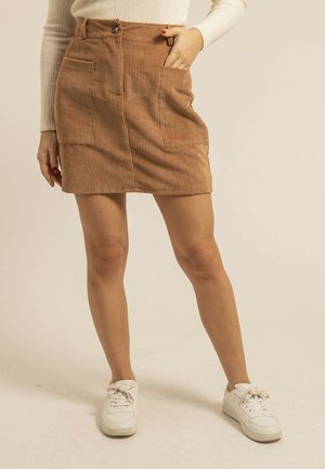 Femme portant une mini-jupe en velours côtelé marron avec des poches avant, des baskets blanches et un haut à manches longues côtelé crème, debout devant un fond uni.