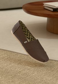 Καφέ καμβάς παπούτσι χωρίς κορδόνια με εσωτερική επένδυση με ζιγκ-ζαγκ σχέδιο, τοποθετημένο σε ένα μπεζ υφαντό χαλί κοντά σε έναν ξύλινο στρογγυλό τραπέζι με βιβλία.