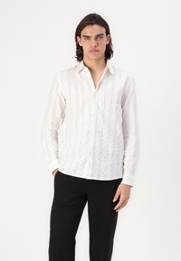 Witte blouse met lange mouwen en verticale blauwe strepen, knoopsluiting aan de voorkant en gestructureerde stof. Gecombineerd met zwarte broek.