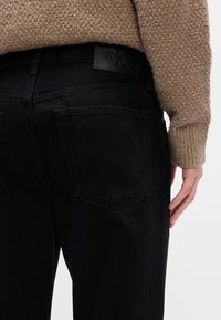 Személy fekete farmernadrágot visel hátsó zsebbel és bézs texturált kötött pulóvert, keze oldalt pihen.