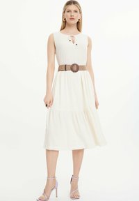 Vestido blanco sin mangas con un escote atado y falda fruncida, combinado con un cinturón marrón trenzado y sandalias de tacón alto de color púrpura pálido.