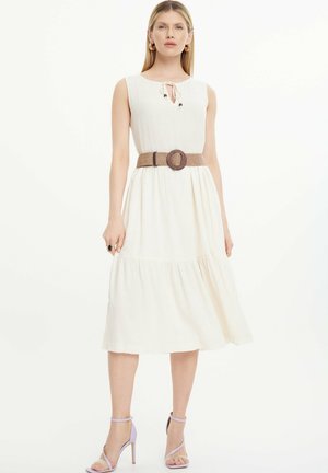 Weißes, ärmelloses Kleid mit einer Krawattenbrust und gerafftem Rock, kombiniert mit einem geflochtenen braunen Gürtel und blasslila High Heels.