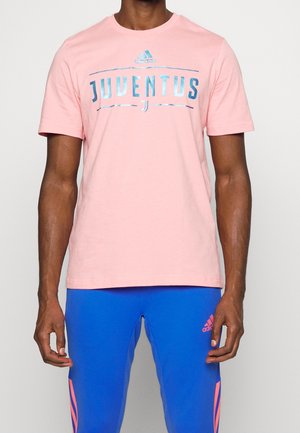 Homme portant un t-shirt Adidas Juventus rose clair et un pantalon Adidas bleu vif avec logo et accents roses, debout devant un fond uni.
