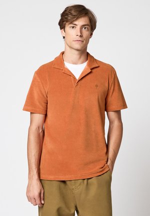 Jeune homme aux cheveux châtain clair portant un polo orange à manches courtes sur un t-shirt blanc et un pantalon kaki, debout avec une main dans la poche.