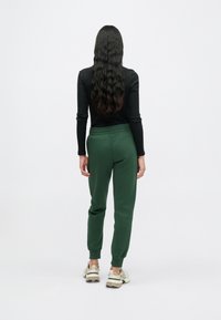 Zaļas sporta bikses ar šaurāku piegriezumu kājām un elastīgu jostasvietu, kopā ar melnu garām piedurknēm kreklu un bēšām sporta kurpēm.