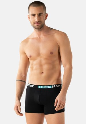 Homme torse nu aux cheveux courts, portant un boxer noir avec la ceinture "ATHENA SPORT", debout devant un fond clair uni.