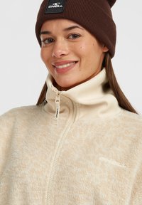 Beige fleecetrui met hoge kraag en ritssluiting, met een gestructureerde oppervlakte en een klein logopatch op de bovenste linkerzijde. Bruine gebreide beanie.