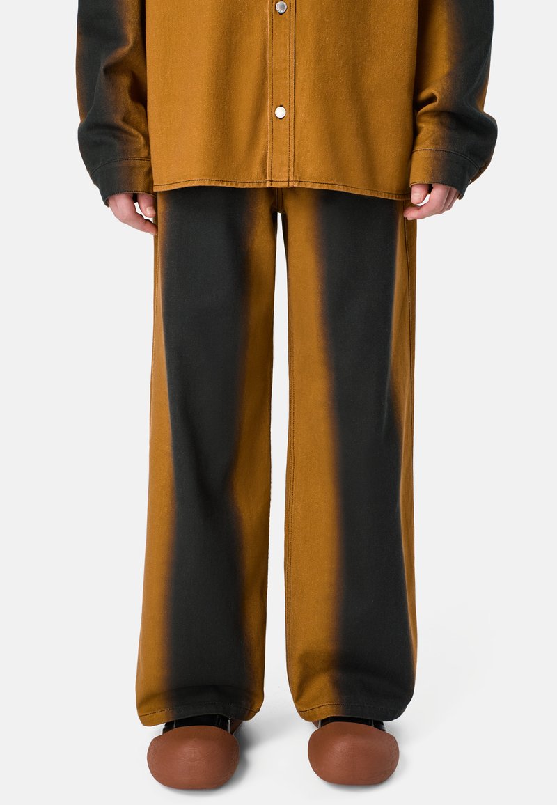 Pantaloni in tessuto marrone e nero con una vestibilità ampia, caratterizzati da cuciture laterali e hardware minimalista, abbinati a scarpe tonde marroni.
