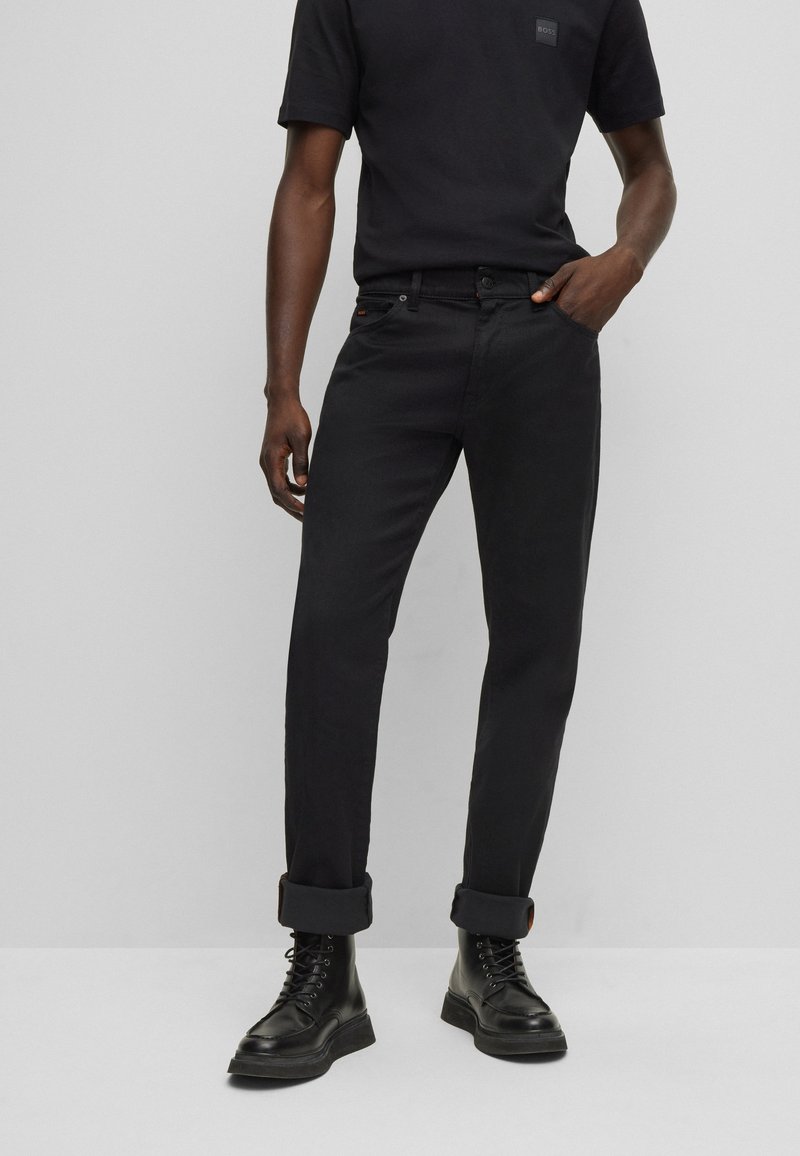 Camisa negra de algodón con un pequeño logo, combinada con jeans negros de corte slim y puños doblados. Se lleva con botas negras con cordones.