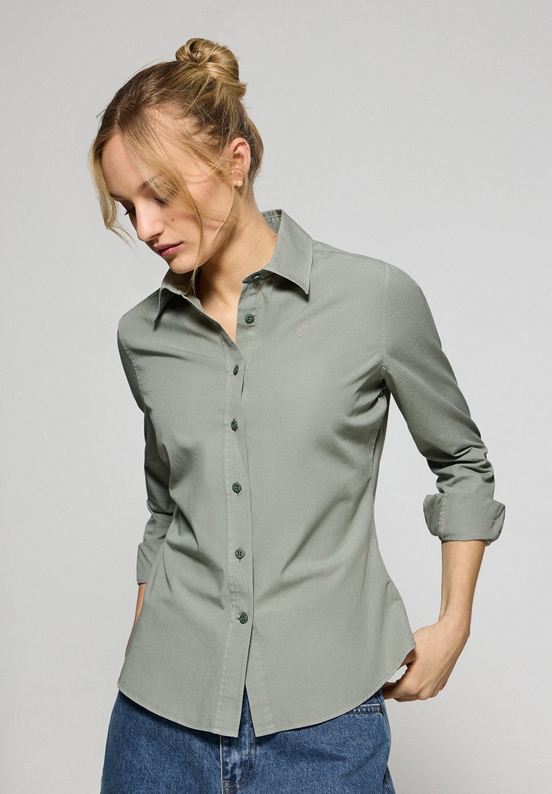 Grön skjorta med knappar, spetsig krage, långa ärmar uppvikt till armbågarna, slät textur och rundad nederkant.