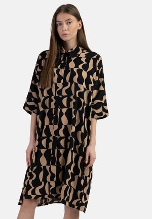 Ung kvinde iført en løs knælængde skjortekjole med sort og beige abstrakt bølget mønster står mod en lys baggrund.