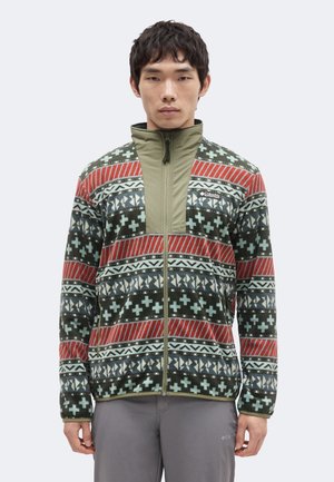 Mann trägt eine Fleecejacke mit Reißverschluss mit grünen, roten, weißen und schwarzen geometrischen und Kreuzmustern, steht vor einem einfarbigen Hintergrund.
