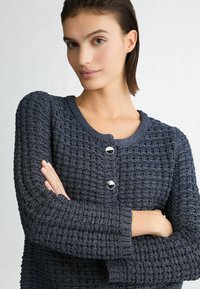 Marineblauwe gebreide cardigan met een gestructureerd patroon, ronde halslijn en zilveren knopen. Voorzien van lange mouwen en een aansluitende silhouette.