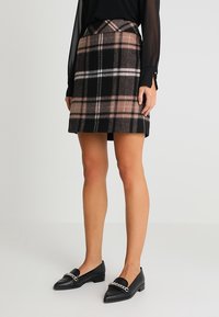 Femme portant un chemisier noir à manches transparentes, une mini-jupe à carreaux marron et noir, et des mocassins noirs pointus avec un détail de chaîne argentée.