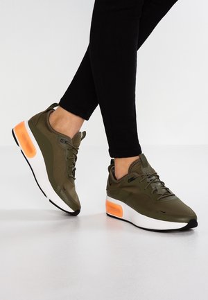 Osoba nosząca oliwkowo-zielone sneakersy z pomarańczowymi poduszkami powietrznymi i czarne spodnie, stojąca na białej podłodze.