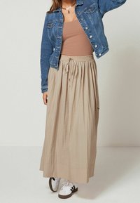 Falda larga beige con cintura elástica y acabado texturizado, combinada con una blusa marrón y una chaqueta vaquera. Zapatillas blancas con detalles negros.