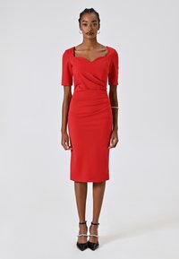 Robe ajustée rouge avec des manches courtes, décolleté carré, détails froncés à la taille et longueur aux genoux. Portée avec des escarpins noirs pointus.