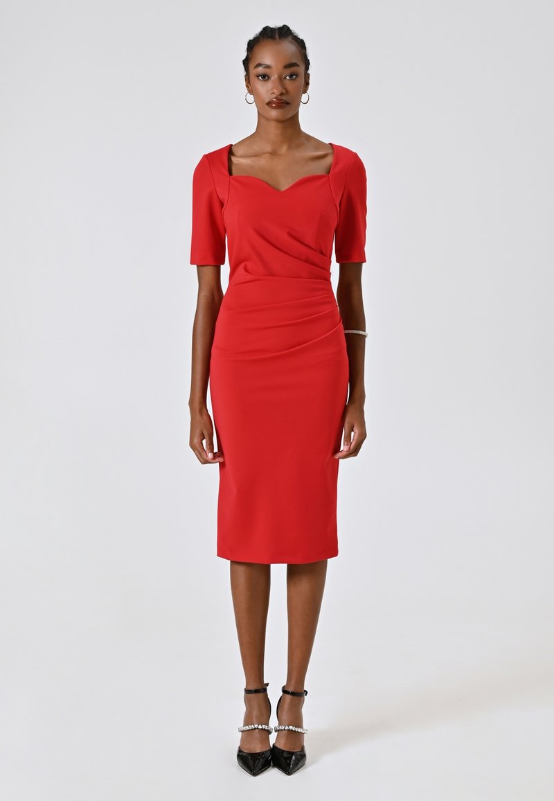 Robe ajustée rouge avec des manches courtes, décolleté carré, détails froncés à la taille et longueur aux genoux. Portée avec des escarpins noirs pointus.