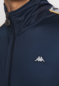 Marinblå zip-up jacka med en slät textur, med en silverdragkedja och en vit logopatch på bröstet. Accentuerad av orange ränder på axlarna.