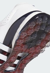 Chaussure de sport blanche avec une tige texturée, des perforations et une semelle extérieure sombre avec des crampons hexagonaux pour la traction. Accentuée par des bandes navy.