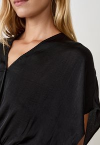 Indumento nero a V, realizzato in tessuto leggero, caratterizzato da una vestibilità ampia, maniche larghe e una superficie liscia, leggermente strutturata.