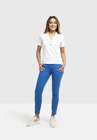 Polo shirt bianco a maniche corte con colletto arricciato, abbinato a pantaloni blue slim-fit e scarpe bianche con accenti crema.