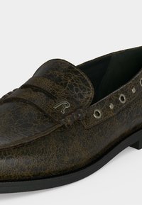 Σκούρο καφέ δερμάτινο μοκασίνι με ραγισμένη υφή, μεταλλικές κλειδαρότρυπες στο πλάι και μικρό ασημένιο διακοσμητικό έμβλημα στο μπροστινό λουρί.