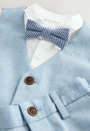 Šviesiai mėlynas džinsinis liemenė su po ja esančia balta palaidine. Papuošta dryžuotu mėlynos ir baltos spalvos petelišku ir rudos spalvos mygtukais ant liemenės.