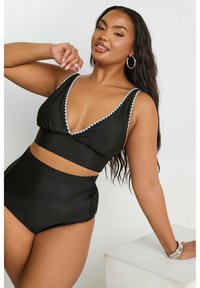 Svart bikini med en djup V-ringad topp och högmidjade bottnar. Har vit scalloped trim och en slät, glänsande tygtextur.