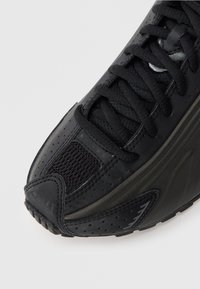 Czarny but sportowy z cholewką z siateczki o perforowanej teksturze, płaskimi sznurowadłami, gumową podeszwą i smukłym designem z subtelnymi akcentami.