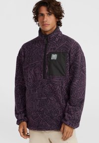 Tumedast lillas fleece-pullover, millel on mustad abstraktseid mustreid, viceeritud esiosa ja tahke must rindkott, millel on logo. Pehme tekstuur.
