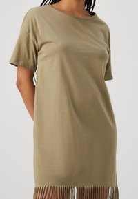 Olivgrön bomulls t-shirt klänning med korta ärmar och en fransad nederkant. Slät textur, lös passform, rund halsringning, avslappnad stil.