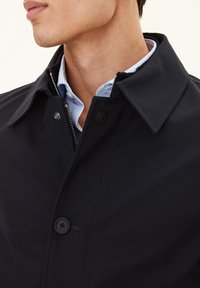 Mörk marinblå trenchcoat med en framträdande krage, enkel knäppning och en dragkedjeficka. Under finns en ljusbl å randig skjorta synlig.