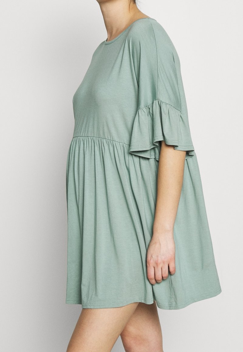 Mujer con un vestido corto y suelto de color verde salvia, con cintura fruncida y mangas cortas con volantes, de pie contra un fondo claro y liso.