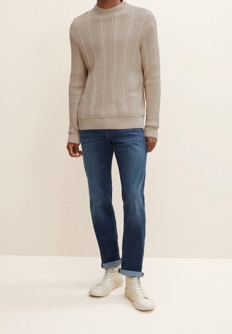 Homme portant un pull beige à torsades, un jean bleu retroussé et des baskets blanches montantes, debout devant un fond clair uni.